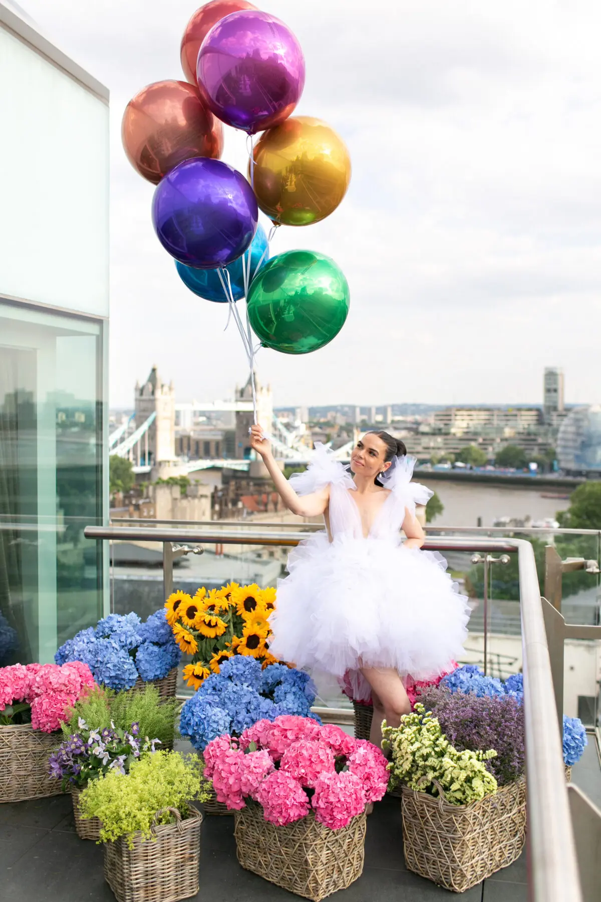 Woman in short white dress surrounded in brightly coloured summer flowers in baskets and holding brightly coloured balloons | Luxury event planner